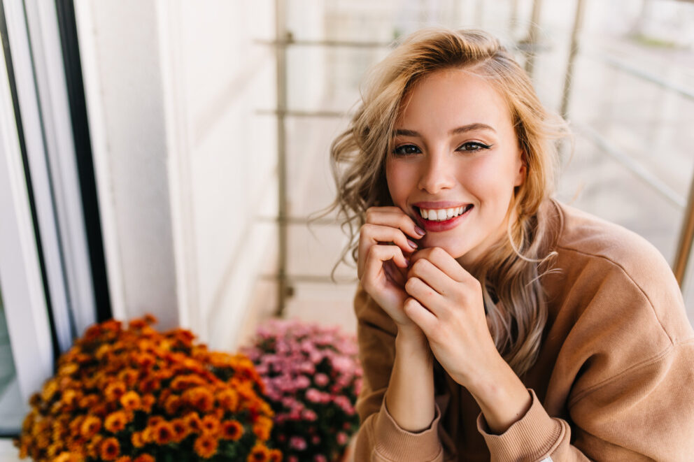 Merry girl with curly hairstyle posing with smile. Elegant female model sitting beside colorful flowers.